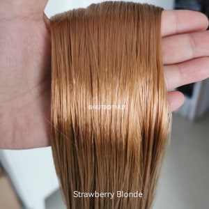 May include: A close-up of a hand holding a bundle of long, straight, strawberry blonde hair extensions. The hair is smooth and shiny, with a warm, reddish-blonde colour.