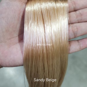 May include: A straight, smooth section of hair extensions in a light sandy beige color. The hair has a glossy, reflective finish and is presented in a vertical orientation. The text @NUTDOTNUT and the words Sandy Beige are printed on the image.