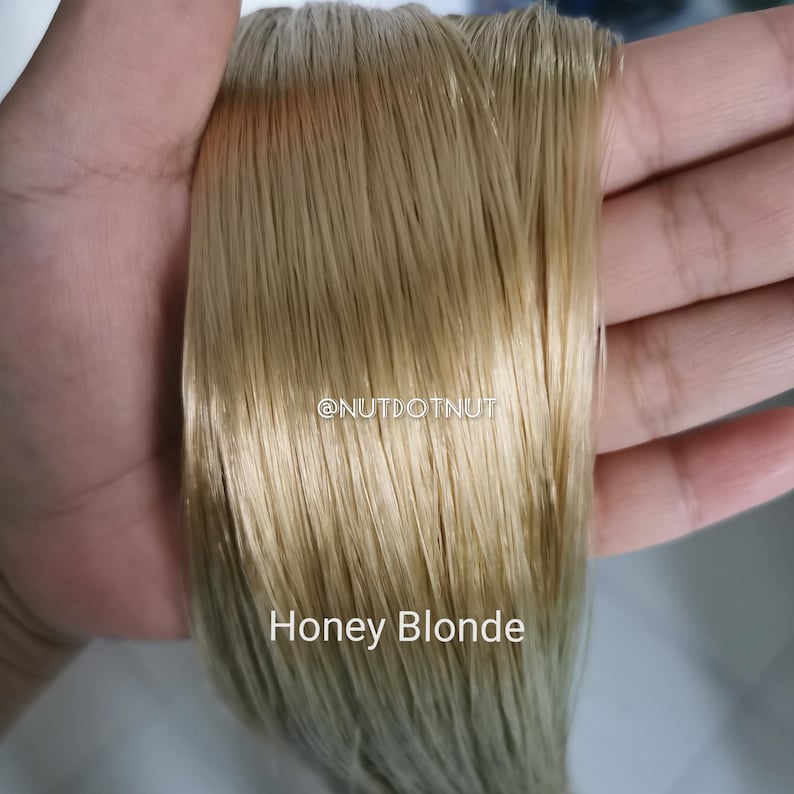 May include: A straight, smooth section of honey blonde hair extensions with a glossy finish. The text @NUTDOTNUT is printed in white in the center, and the words Honey Blonde are printed in white at the bottom of the hair strands.