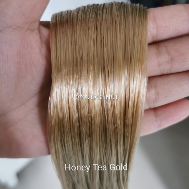 May include: A straight, honey-blonde hair extension piece with a smooth, glossy texture. The text @NUTDOTNUT is printed in the center, and the text Honey Tea Gold is printed at the bottom. The hair is light golden brown with subtle highlights.