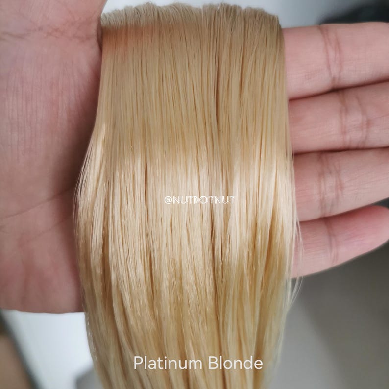 May include: A straight, smooth section of hair extensions in a light platinum blonde shade. The hair strands are arranged vertically and appear sleek with a glossy finish. The text @NUTDOTNUT and the words Platinum Blonde are visible on the image.