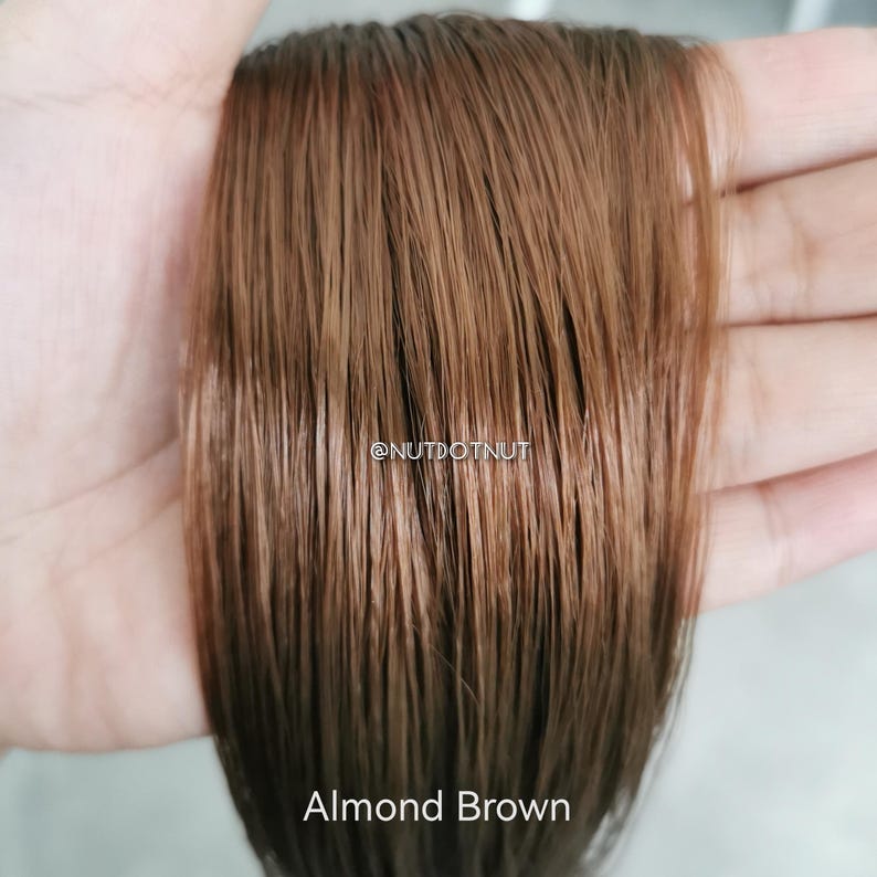 May include: Close-up of a hand holding a section of straight, brown human hair. The hair is a light brown color with a slight reddish hue. The text "Almond Brown" is visible at the bottom of the image.