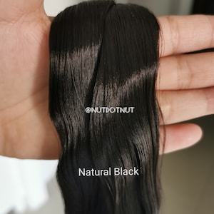 May include: A close-up of a long, straight, black hair extension. The hair is smooth and shiny, and it is held in a hand. The text "@NUTDOTNUT" and "Natural Black" is visible in the image.