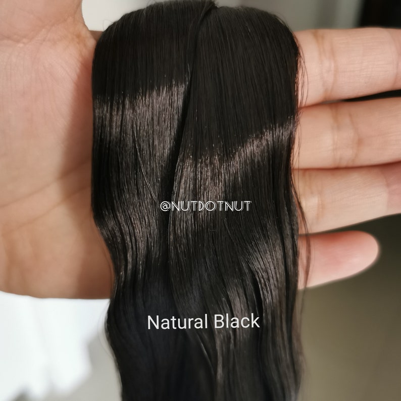 May include: A close-up of a long, straight, black hair extension. The hair is smooth and shiny, and it is held in a hand. The text "@NUTDOTNUT" and "Natural Black" is visible in the image.