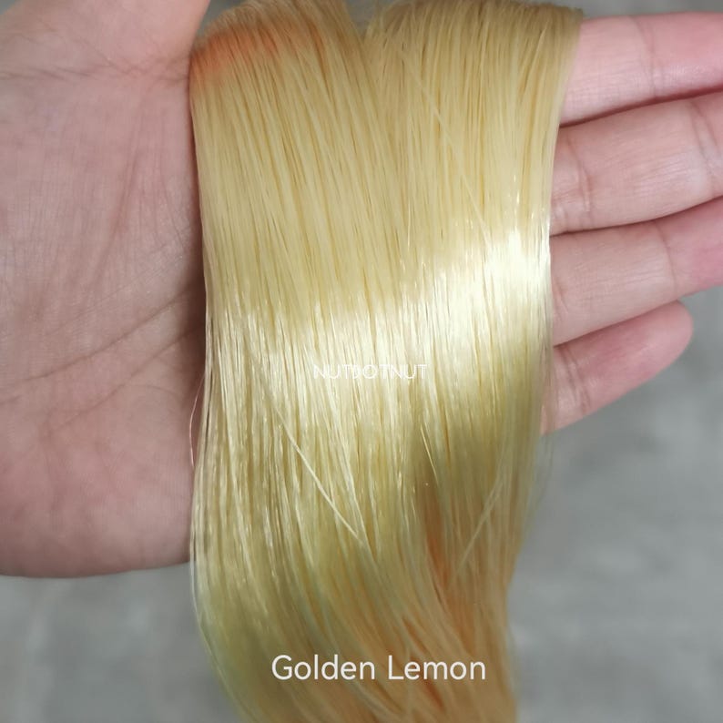 May include: A bundle of straight, pale yellow synthetic hair fibers with a smooth, glossy texture. The fibers are light, translucent, and have a consistent golden lemon color throughout. The text Golden Lemon is printed at the bottom.