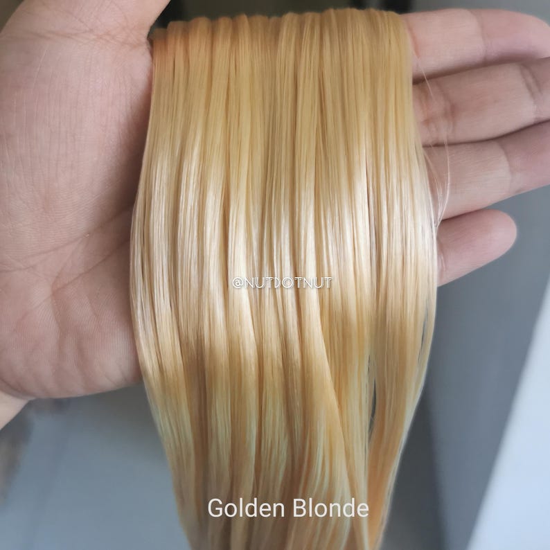 May include: A straight, smooth section of synthetic hair extensions in a light golden blonde shade. The hair strands are uniform in color and texture, featuring the text @NUTDOTNUT printed in the center and Golden Blonde at the bottom.