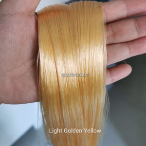 May include: A straight, light golden yellow hair extension piece with a smooth, glossy texture. The text @NUTDOTNUT is visible in the center, and the text Light Golden Yellow is printed at the bottom of the straight, synthetic-looking strands.