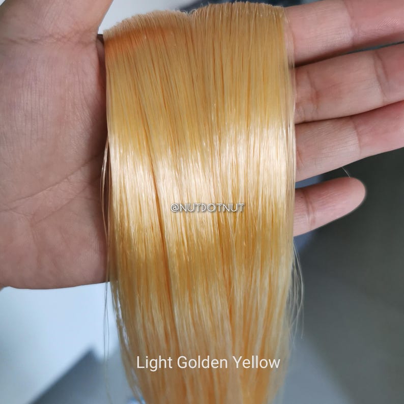 May include: A straight, light golden yellow hair extension piece with a smooth, glossy texture. The text @NUTDOTNUT is visible in the center, and the text Light Golden Yellow is printed at the bottom of the straight, synthetic-looking strands.