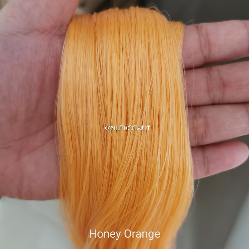 May include: A bundle of straight, smooth synthetic hair fibers in a vibrant honey orange color. The fibers are fine and have a slight sheen. The text @NUTDOTNUT is visible in the center, and the text Honey Orange is printed at the bottom.