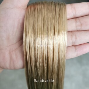 May include: A straight, light golden-blonde hair extension piece with a smooth, glossy texture. The text @NUTDOTNUT is printed in white in the center, and the word Sandcastle is printed in white at the bottom of the hair strand.