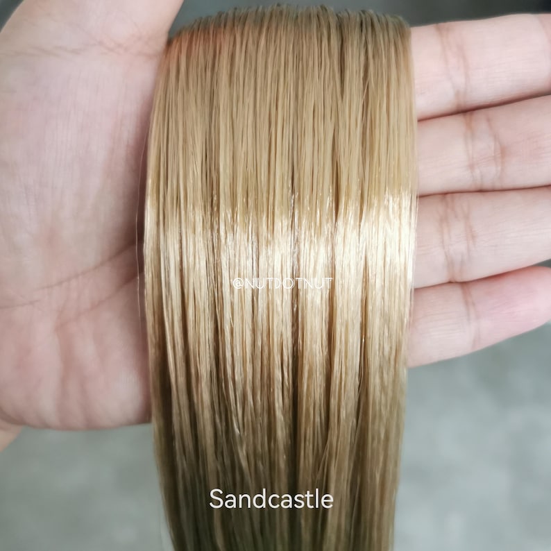 May include: A straight, light golden-blonde hair extension piece with a smooth, glossy texture. The text @NUTDOTNUT is printed in white in the center, and the word Sandcastle is printed in white at the bottom of the hair strand.