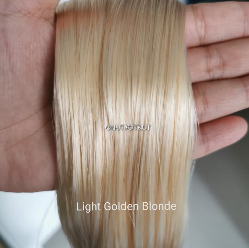 May include: A straight, light golden blonde hair extension piece featuring a smooth, silky texture. The text @NUTDOTNUT is printed in the center, and the words Light Golden Blonde are printed at the bottom of the hair strands.