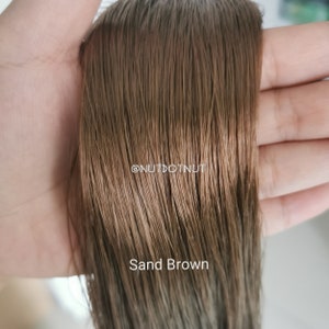 May include: A close-up of a strand of light brown human hair, showing the texture and color. The hair is labeled "Sand Brown" and the text "@nutdotnut" is visible in the image.