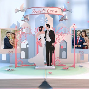 May include: A paper pop-up card with a wedding theme. The card features a couple in a white dress and black suit standing in front of a castle. The card also includes two photos of the couple, one on each side of the castle. The card is decorated with pink and blue flowers and hearts. The text "Anna & David" is written on the top of the card.