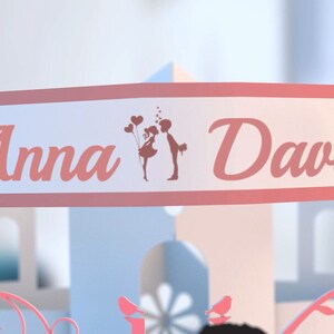 May include: A white banner with pink trim and the names "Anna" and "David" in pink script. A silhouette of a couple holding balloons and kissing is in the center of the banner.