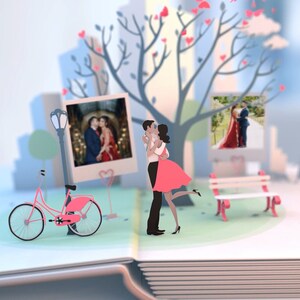 May include: A pop-up book with a papercraft scene of a couple kissing under a tree with pink hearts. There is a pink bicycle, a bench, and three instant photos of the couple.