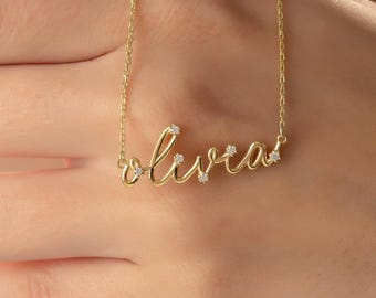 May include: A gold-colored necklace with the name "Olivia" in a cursive font. The necklace has small, clear gemstones embedded in the letters. The chain is delicate and thin, and the necklace is displayed on a person's hand.