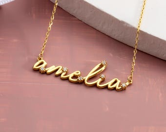 May include: A gold-colored necklace with the name "amelia" in cursive script. The necklace has small, clear gemstones embedded in the letters. The chain is delicate, and the necklace is displayed on a pink surface.