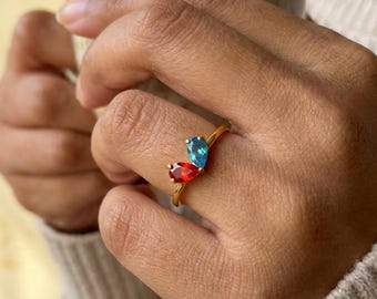 May include: A gold ring featuring two pear-shaped gemstones, one vibrant red and one bright blue, set in a bypass style. The band is thin and polished, showcasing the contrasting colors of the teardrop-cut stones positioned side by side on the finger.