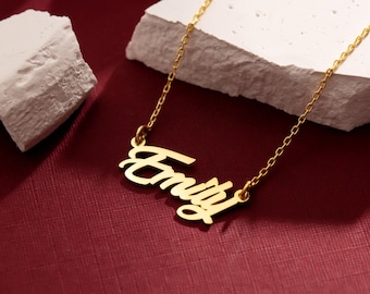 May include: A gold-colored necklace with the name "Emily" in a cursive font. The pendant hangs from a delicate chain. The necklace is displayed on a white, textured surface against a deep red background.