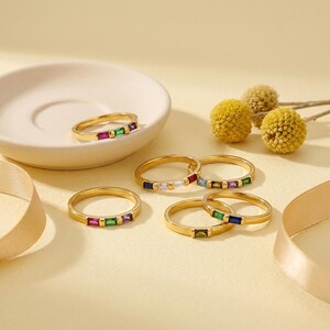 May include: Five gold rings, each featuring a row of three small, rectangular, multi-colored gemstones. The stones include shades of red, green, blue, and clear white. One ring rests inside a small, round, off-white ceramic dish.