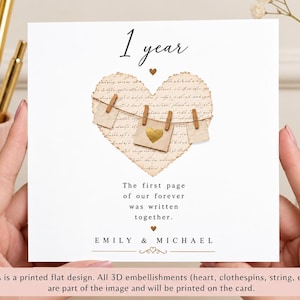 May include: A square greeting card featuring the text 1 year at the top and a central heart shape with vintage script. A string with three small paper squares and wooden clothespins is printed across the heart. The text reads The first page of our forever was written together, followed by the names EMILY & MICHAEL.