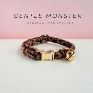 May include: A leopard print pet collar with a gold-colored buckle and a small bell. The text "GENTLE MONSTER PERSONALIZED COLLARS" is displayed above the collar.