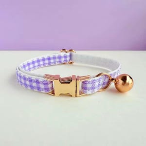 Puede incluir: Un collar para gato con un estampado de cuadros vichy en lavanda y blanco. El collar tiene una hebilla de metal dorado y una campana a juego. El fondo es de color púrpura suave.