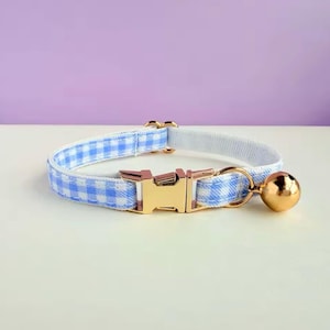 Puede incluir: Un collar para gato a cuadros azules y blancos con una hebilla dorada y una pequeña campana dorada. El collar es de tela y tiene un interior blanco. El fondo es de color lila claro.