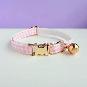 Puede incluir: Un collar para gatos a cuadros rosas y blancos con una hebilla de metal dorada y una campana a juego. El collar está hecho de una tela suave. El collar mide aproximadamente 25 cm de largo.