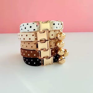Peut inclure: Une pile de cinq colliers pour chats de différentes couleurs, dont blanc à pois noirs, beige à pois marron et marron à pois blancs. Chaque collier est doté d'une boucle en métal doré et d'un charm en forme de cloche doré assorti.