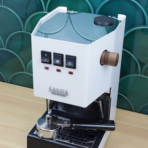 May include: A white Gaggia espresso machine with a black base and a wooden handle. The machine has three illuminated buttons and a silver portafilter. The Gaggia logo is visible on the front.
