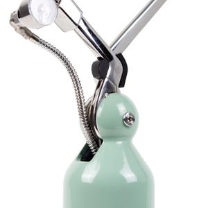 May include: A close-up of a mint green and silver metal lamp. The lamp has a rounded base, a flexible silver hose, and a gauge. The lamp's design is modern and industrial, with a focus on functionality and style.