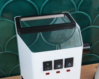 May include: White espresso machine with a black and silver top and three buttons. The buttons are labeled with icons for on, coffee cup, and steam. The machine has the text "Gaggia Classic" on the front.