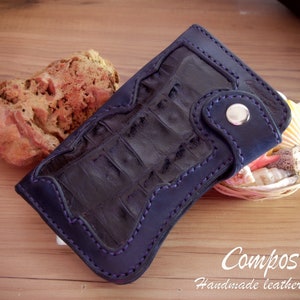 May include: A dark blue leather wallet with a crocodile pattern and a stitched border. The wallet has a snap closure and a small pocket on the front. The wallet is lying on a wooden surface.