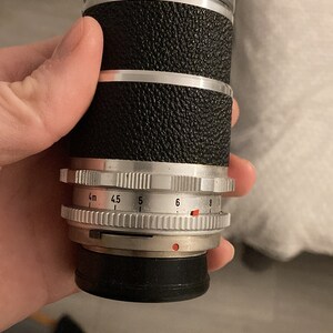 May include: A silver and black camera lens with a depth of field scale marked in meters and f-stops. The lens has a rubberized grip and a silver ring with a red dot.