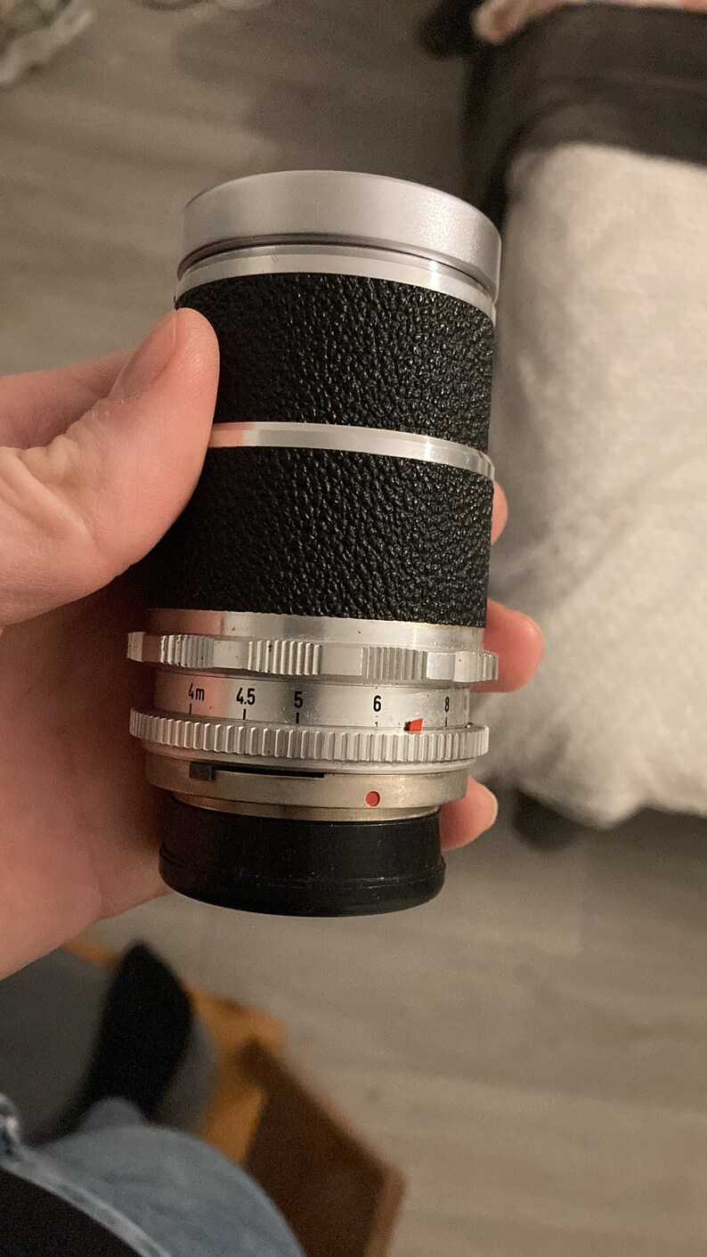 May include: A silver and black camera lens with a depth of field scale marked in meters and f-stops. The lens has a rubberized grip and a silver ring with a red dot.