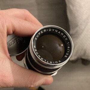 May include: A black and silver camera lens with the text "Voigtlander Super-Dynarex 1:4/135" printed around the edge.