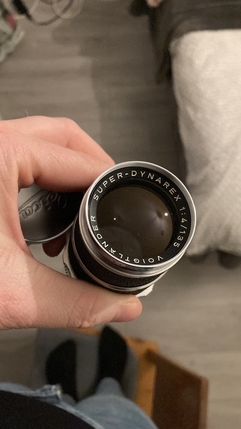 May include: A black and silver camera lens with the text "Voigtlander Super-Dynarex 1:4/135" printed around the edge.