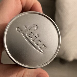 May include: A silver lens cap with the word "Leica" embossed on it.