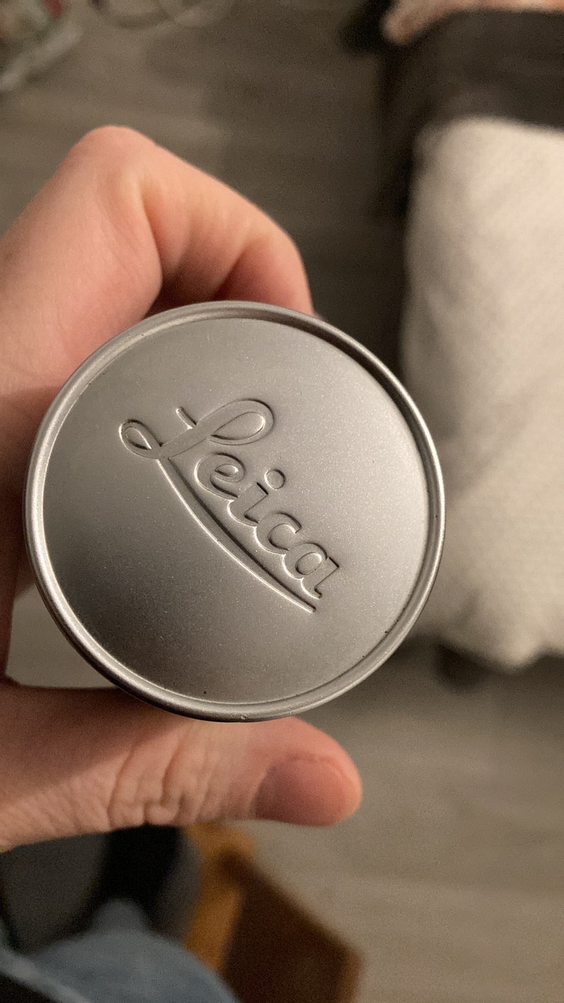 May include: A silver lens cap with the word "Leica" embossed on it.