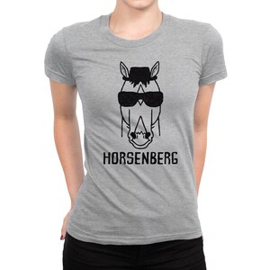 Może przedstawiać: Szary T-shirt z czarną ilustracją konia w okularach przeciwsłonecznych i kapeluszu. Pod koniem nadrukowany jest tekst "HORSENBERG".