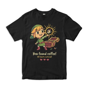 Puede incluir: Una camiseta negra con una imagen de dibujos animados de Link de la serie de videojuegos The Legend of Zelda. Link sostiene una taza de café y está de pie frente a un cofre del tesoro abierto. El texto en la camiseta dice "You found coffee! All hearts restored!" con tres corazones rojos debajo del texto.