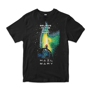 Könnte beinhalten: Schwarzes T-Shirt mit einem grafischen Design. Es zeigt eine Figur auf einer Struktur, die von einem gelben Licht beleuchtet wird. Der Text "BELIEVE IN THE HAIL MARY" und "PROJECT HAIL MARY" ist weiß gedruckt.