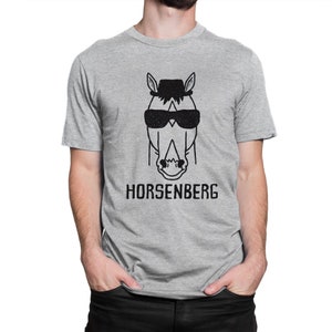 Może przedstawiać: Szary T-shirt z czarno-białym rysunkiem konia w okularach przeciwsłonecznych i kapeluszu. Pod rysunkiem znajduje się napis "HORSENBERG".