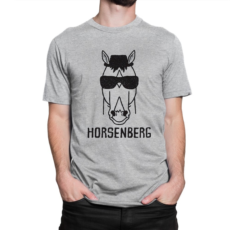 Może przedstawiać: Szary T-shirt z czarno-białym rysunkiem konia w okularach przeciwsłonecznych i kapeluszu. Pod rysunkiem znajduje się napis "HORSENBERG".