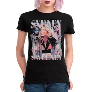 May include: Black t-shirt featuring a graphic of Sydney Sweeney with the text "SYDNEY SWEENEY" in a retro style. The design includes multiple images of the actress in various poses, with a pink and blue color scheme.