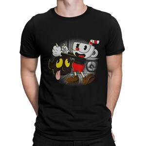 Peut inclure: T-shirt noir avec une image de dessin animé d'un personnage du jeu vidéo Cuphead. Le personnage tient un chat noir avec un sourire diabolique. Le texte "The Devil's Advocate" est imprimé sur le t-shirt.