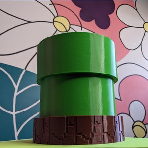 May include: A green and brown 3D printed pipe, resembling a video game item, is sitting on a green surface. The pipe is made of two sections, with a brown base.