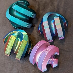 May include: Four abstract, iridescent objects with a curved, layered design. The colors shift from teal to purple and pink, creating a dynamic visual effect. The objects appear to be 3D-printed, with visible layer lines.